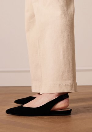 Zwarte fluwelen slingback balletjes met een spitse neus, gedragen met beige wijde broekspijpen, die een gladde textuur en minimalistisch ontwerp tonen.