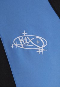 Blåt stof med en glat tekstur, prydet med et hvidt broderet logo med stjerner og en oval form, kontrasteret af sorte accenter.