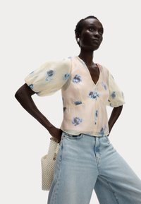 Blusa floreale con maniche a palloncino, tessuto trasparente in un delicato rosa con fiori blu, abbinata a jeans rilassati blu chiaro e una borsa con texture a perle.