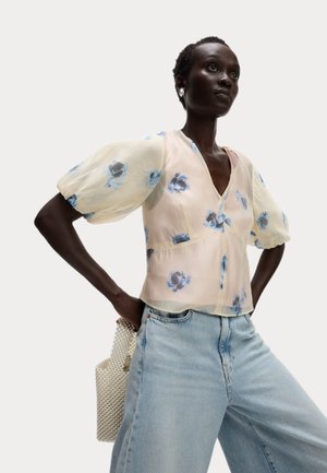 Blouse florale à manches bouffantes, en tissu transparent de rose pâle avec des fleurs bleues, associée à un jean décontracté bleu clair et un sac à texture perle.