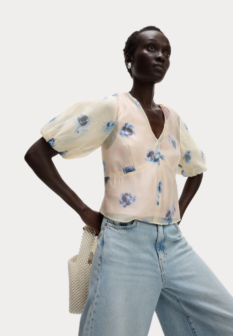 Blusa floreale con maniche a palloncino, tessuto trasparente in un delicato rosa con fiori blu, abbinata a jeans rilassati blu chiaro e una borsa con texture a perle.