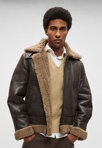 Barna bőrdzseki műszőrméből készült gallérral és mandzsettával. Két elülső zsebbel és cipzáras lezárással rendelkezik. Bézs pulóverre viselve.