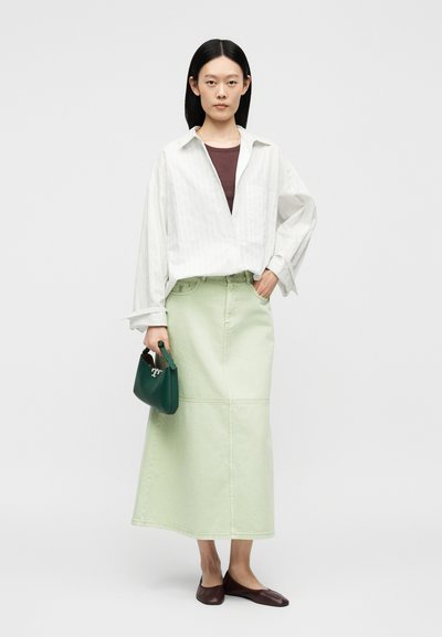 Femme aux cheveux noirs et raides portant une chemise blanche ample par-dessus un haut marron, une jupe midi vert clair, des ballerines marron, tenant un petit sac à main vert foncé.