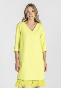 Blondynka w żółtej sukience z dekoltem w literę V, z rękawami o długości trzech czwartych i piórkowym wykończeniem u dołu, stojąca na jednolitym tle.