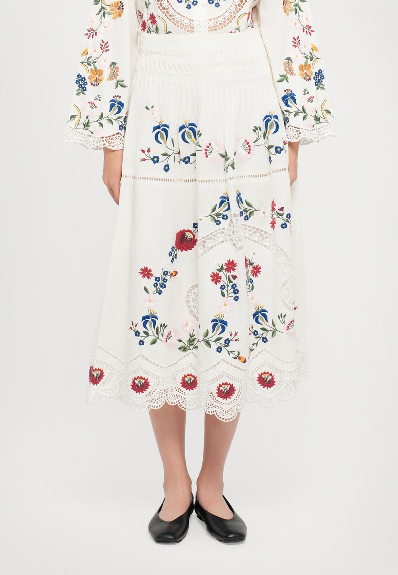 Weißer bestickter Rock mit bunten Blumenmustern, gewelltem Spitzenrand und tailliertem Bund. Kombiniert mit schwarzen Ballerinas.