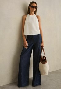 Biała bezrękawnikowa bluzka z wysokim dekoltem, zestawiona z szerokimi spodniami z ciemnego denimu. Do stroju dodano beżowo-brązową torbę typu tote.