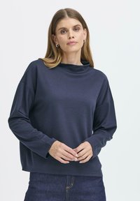 Mörkblå långärmad tröja med hög krage och avslappnad passform. Mjuk textur och vida ärmar, kombinerat med mörkblå denimjeans.