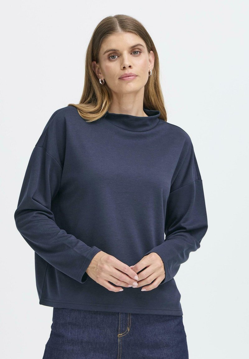 Mörkblå långärmad tröja med hög krage och avslappnad passform. Mjuk textur och vida ärmar, kombinerat med mörkblå denimjeans.