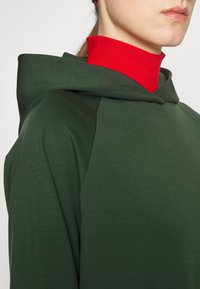 Felpa verde con tessuto a trama, con uno strato rosso a collo alto sotto e un cappuccio prominente. Design semplice, senza hardware visibile.