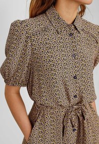 Camicia a stampa leopardata in tessuto leggero; presenta un collo a camicia, maniche corte a sbuffo, bottoni frontali e un dettaglio con vita annodata.