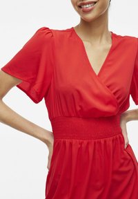Červené šaty vyrobené z ľahkej tkaniny, s hlbokým výstrihom do V, krátkymi rukávmi, smockovaným pásom a jemnými plisovaním na sukni.