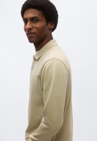 Világos bézs, hosszú ujjú galléros póló sima textúrával és laza szabással, oldalnézetből ábrázolva.