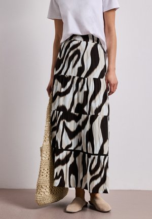 Femme portant une jupe à volants imprimée zèbre noire et blanche, un haut blanc à manches courtes, des chaussures beige à enfiler, tenant un sac en paille tressée.