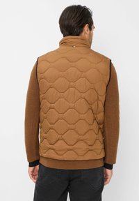 Gilet trapuntato marrone con motivi esagonali, collo alto e senza maniche. Indossato sopra una camicia a maniche lunghe marrone, con dettagli neri.