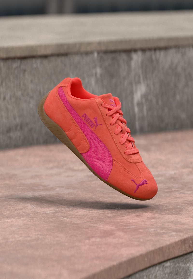 Baskets Puma orange et rose avec semelle marron, flottant au-dessus d'une surface en pierre avec un mur de pierre gris en arrière-plan.