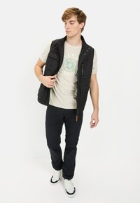 Schwarze ärmellose Jacke über beigem T-Shirt mit grünem Druck, kombiniert mit schwarzen Hosen und weißen Sneakers mit schwarzen Akzenten.