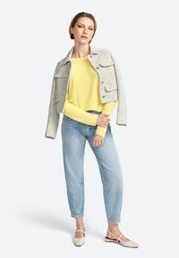 Helle graue Tweedjacke mit zwei Vorderaschen über einem gelben Langarmoberteil, kombiniert mit hellblauen Jeans und beigen Riemensandalen.