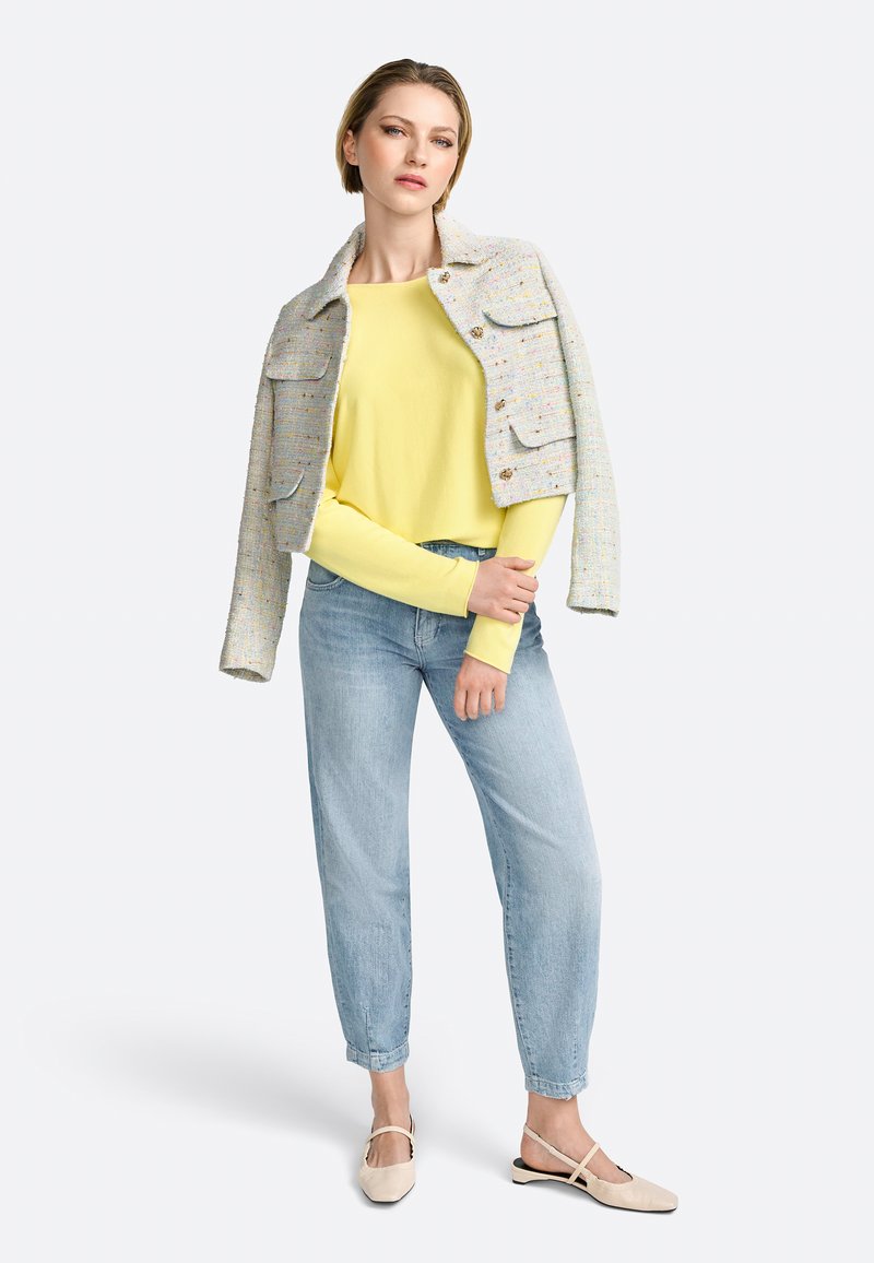 Helle graue Tweedjacke mit zwei Vorderaschen über einem gelben Langarmoberteil, kombiniert mit hellblauen Jeans und beigen Riemensandalen.