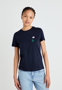 Marineblå kortermet T-skjorte i bomull, med en liten rosa blomsterbrodering på venstre brystområde, kombinert med lyseblå jeans.