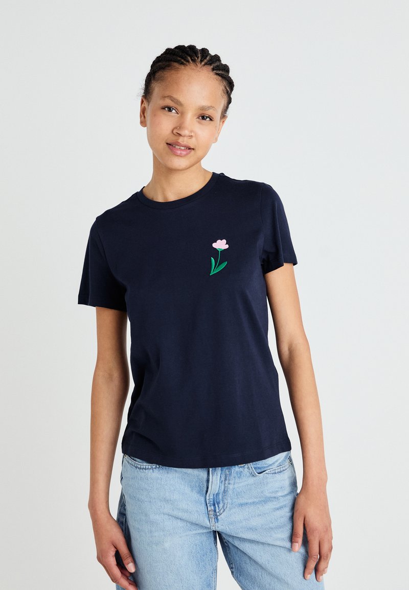 Marineblå kortermet T-skjorte i bomull, med en liten rosa blomsterbrodering på venstre brystområde, kombinert med lyseblå jeans.