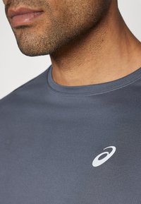 Graues Sporthemd mit rundem Ausschnitt, kurzen Ärmeln und einem dezenten weißen Logo im Brustbereich. Glatte Stofftextur.