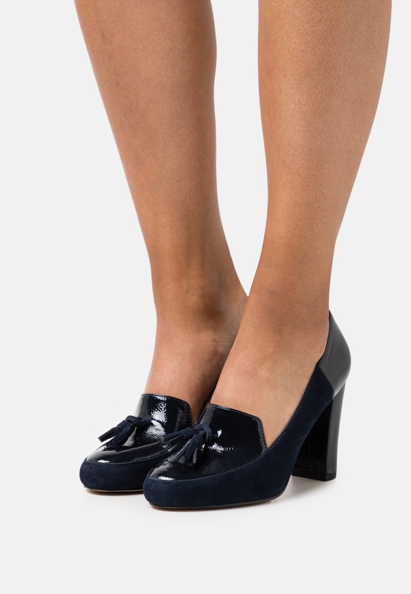 Femme portant des mocassins à talons hauts en cuir verni bleu marine et en daim, ornés de détails à glands, debout sur une surface gris clair.