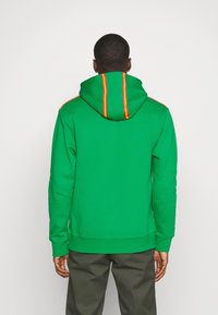 Sudadera verde con capucha, con rayas naranjas a lo largo de la espalda y los hombros. El material parece suave; el diseño es casual y relajado.