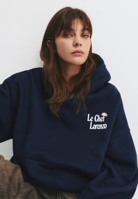 Navy modrý mikina vyrobená z bavlny, s přední kapsou a vyšitým textem "Le Chef Lorenzo" s malým designem houby.