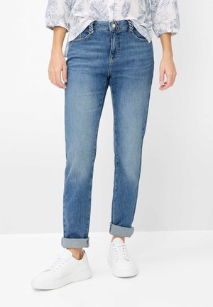 Kvinde iført optrukne blå cowboybukser, hvide sneakers og en hvid blomsterbluse med opsmøgede ærmer, stående mod en hvid baggrund.