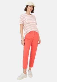 Camicia a righe rossa e bianca, pantaloni corti corallo e sneakers bianche. Il modello indossa un cappello beige chiaro e accessori. Outfit casual.