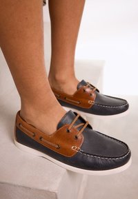 Chaussures mocassins bicolores avec un corps en cuir bleu foncé, des accents en cuir marron, des détails de laçage et une semelle en caoutchouc blanche.
