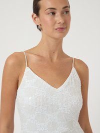 Γυναίκα που φοράει ένα λευκό φόρεμα με χρυσές παγιέτες και λεπτές τιράντες, κοιτάζοντας ελαφρώς προς τα δεξιά, με ασημένια σκουλαρίκια κρίκους και φυσικό μακιγιάζ.