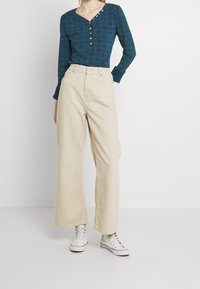 Tyrkysový vzorovaný dlouhý rukáv top s detaily knoflíků; spárováno s krémovými kalhotami s širokými nohavicemi a bílými teniskami, s uvolněným střihem.