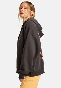 Ciemnoszary hoodie z przednią kieszenią typu kangur, luźny krój i ściągacze przy mankietach. Zawiera nadrukowany tekst i grafiki w kontrastujących kolorach.