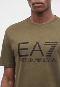 Κοντινό ενός ατόμου που φοράει ελιάς πράσινη μπλούζα με μαύρο κείμενο και λογότυπο "EA7 Emporio Armani" στο στήθος.