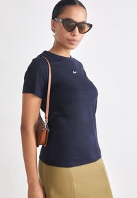 Námornícky modré bavlnené tričko s krátkymi rukávmi, na hrudi s malým logom. V kombinácii s priliehavou béžovou sukňou a hnedou crossbody taškou.