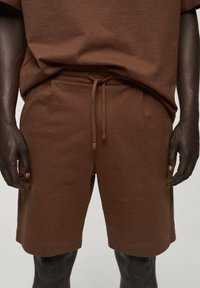 Personne à la peau foncée portant un short en maille marron avec cordon de serrage et un haut marron assorti, se tenant avec les mains détendues sur les côtés.