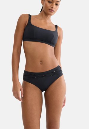 Frau trägt einen schwarzen Bikini mit asymmetrischem Oberteil und hochgeschnittenem Unterteil mit Schnürungsdetails und kleinen goldenen Perlen.