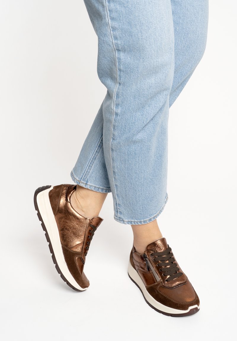 Personne portant un jean bleu clair et des baskets marron métallisé avec des semelles blanches et des lacets noirs, debout sur un fond blanc.