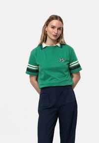 T-shirt en coton vert avec un col rayé blanc et bleu, des rayures blanches sur les manches, et le numéro "75" en noir sur le devant.