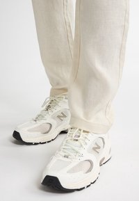 Baskets New Balance blanches et beiges portées avec un pantalon beige clair sur fond blanc.