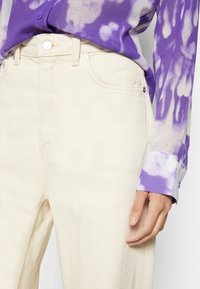 Jeans de ganga branca com um acabamento texturizado, combinados com uma camisola de manga longa em tie-dye roxo, com punhos suaves e botões.