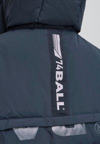 Chaqueta acolchada de tela azul marino oscuro con una textura suave. Presenta una gran etiqueta de logo vertical en blanco en la parte trasera y detalles impresos.