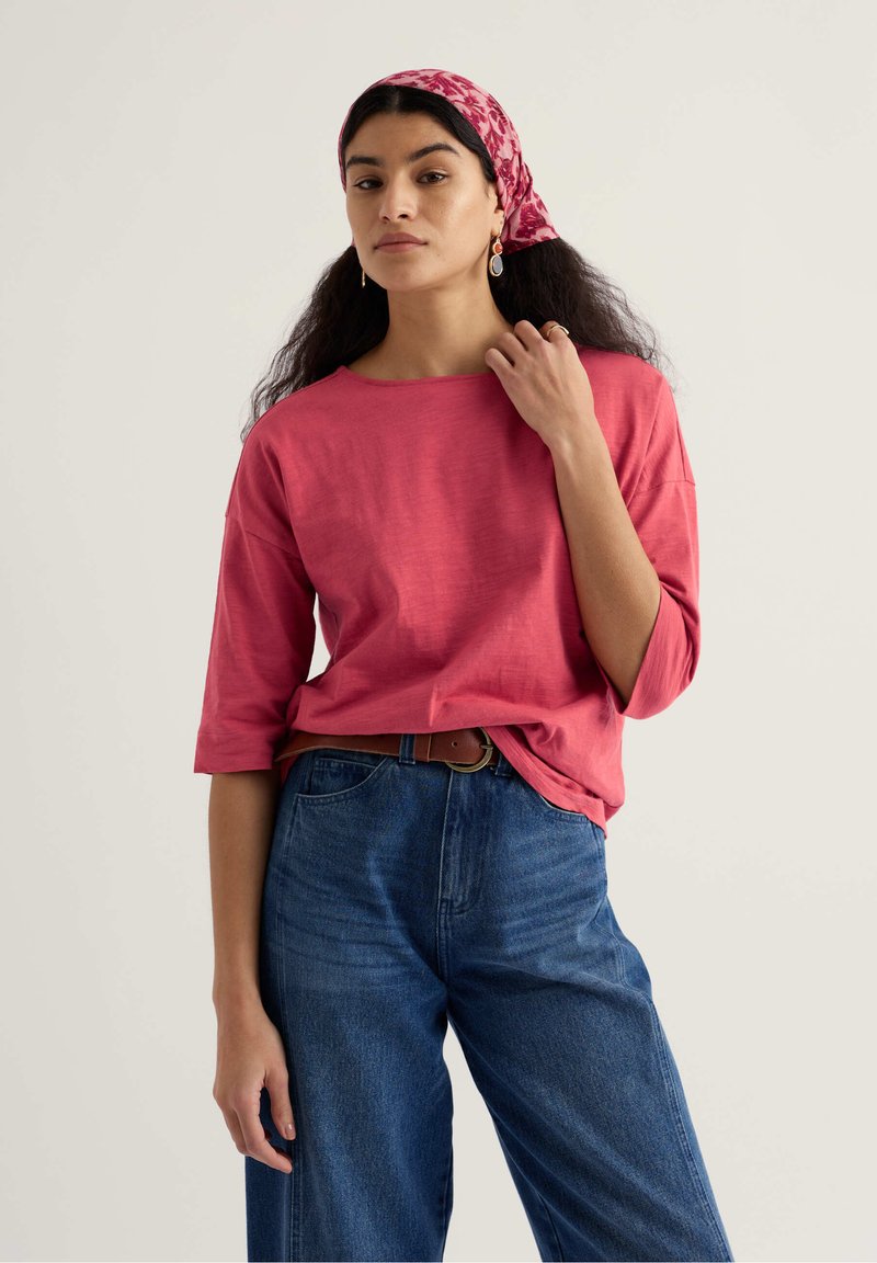 Femme portant un haut rose à manches trois-quarts, un jean taille haute bleu, une ceinture marron et un foulard rouge à motifs floraux, debout devant un fond uni.