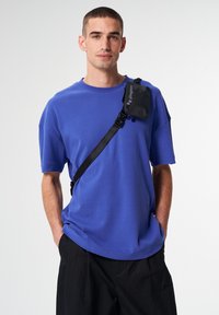 Blaues, oversized T-Shirt mit kurzen Ärmeln und rundem Ausschnitt, kombiniert mit lockeren schwarzen Hosen und einer kleinen schwarzen Umhängetasche.