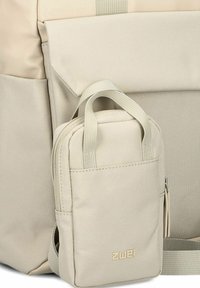Mochila beige hecha de tela texturizada, con un pequeño bolsillo desmontable con asas y el logo de la marca visible en la parte delantera.