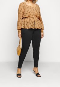 Světle hnědá halenka s puntíky, peplum pasem, dlouhými rukávy a knoflíkovými detaily, kombinovaná s černými slim-fit legínami a taškou přes rameno.