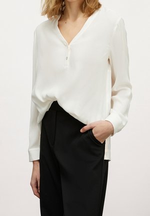 Femme portant un chemisier blanc à manches longues boutonné, rentrée dans un pantalon noir taille haute, avec une main dans une poche.