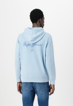 Sweatshirt azul claro feito de tecido macio, com capuz e design de logotipo nas costas. Usado com jeans azuis em um ambiente casual.