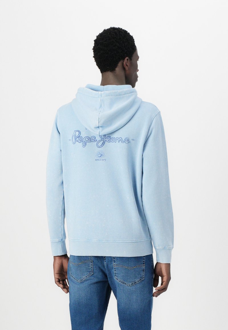 Sweatshirt azul claro feito de tecido macio, com capuz e design de logotipo nas costas. Usado com jeans azuis em um ambiente casual.